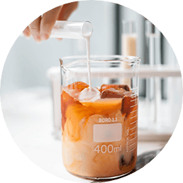 Coffee in a beaker with ice and a hand pouring cream from a test tube into the beaker