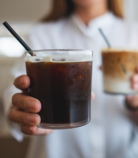 One person holding two glasses of iced coffee, each with a straw in the glass. One glass is black coffee, ther other is layered with cream